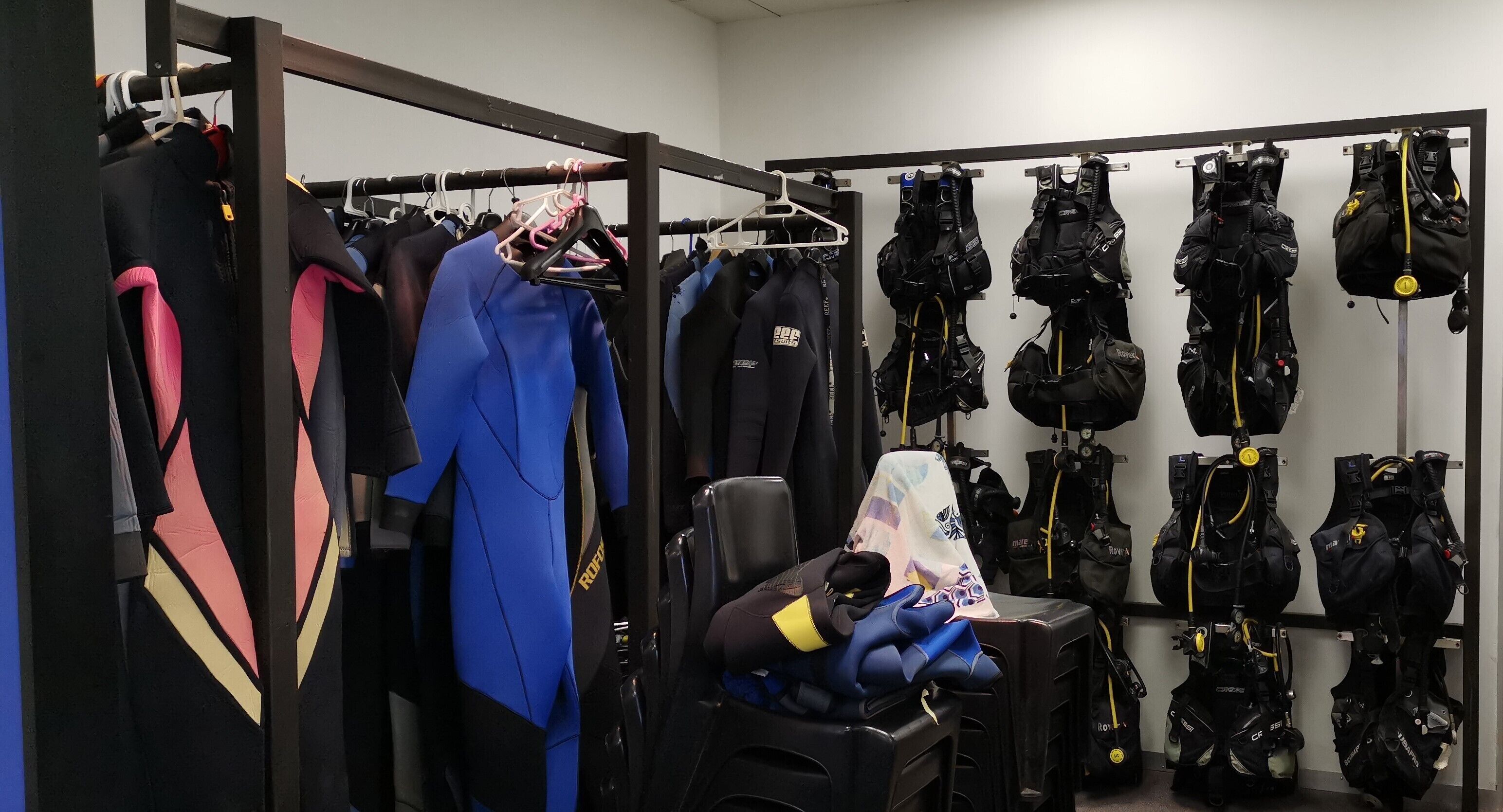 A storage room with rows of black diving gear, including wetsuits and backpacks, and a chair with blue gloves on it.