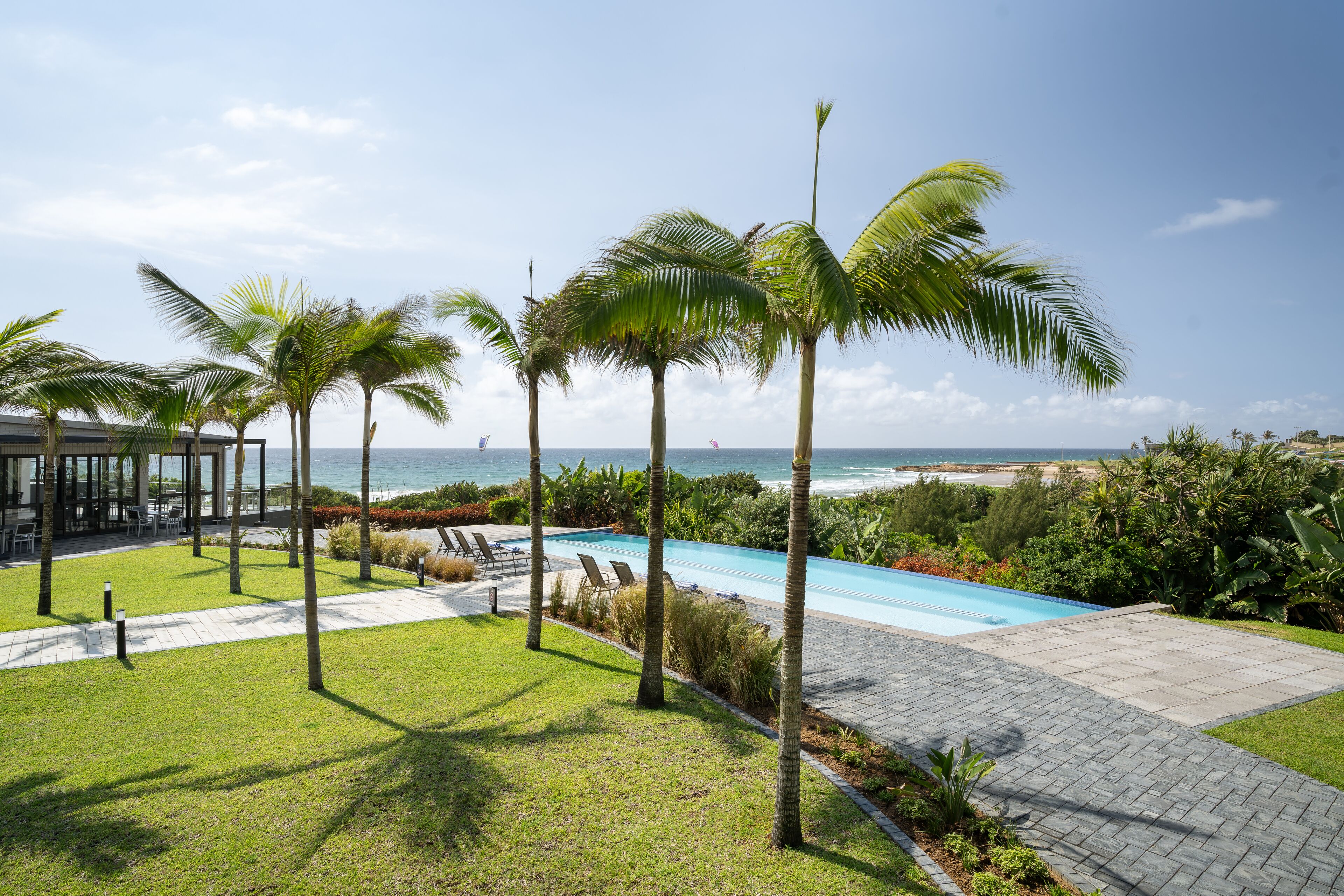 A pool area with palm trees, a view of the ocean, and a patio with chairs.