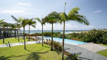 A pool area with palm trees, a view of the ocean, and a patio with chairs.