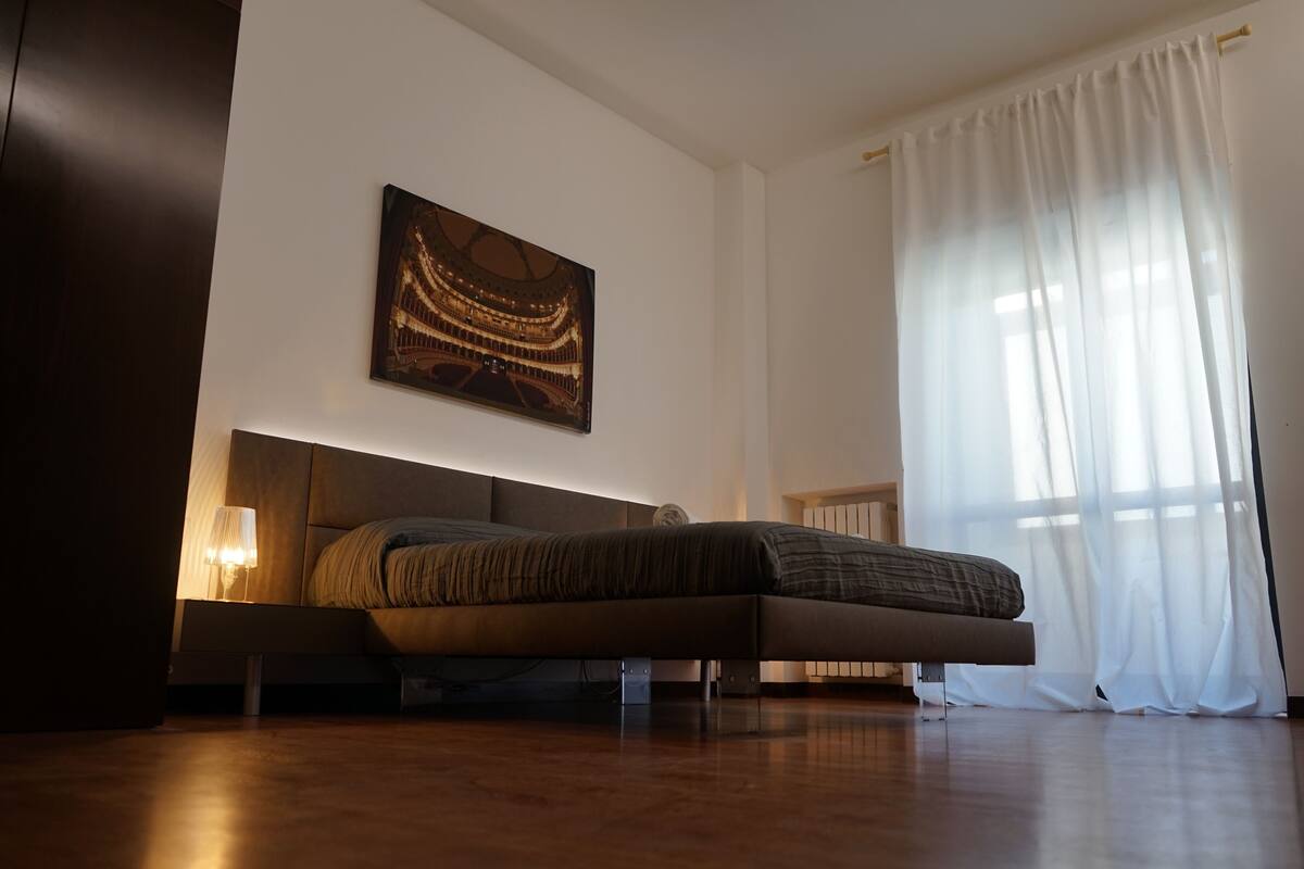 A modern bedroom with a bed, a wall-mounted artwork, and a large window with sheer curtains.