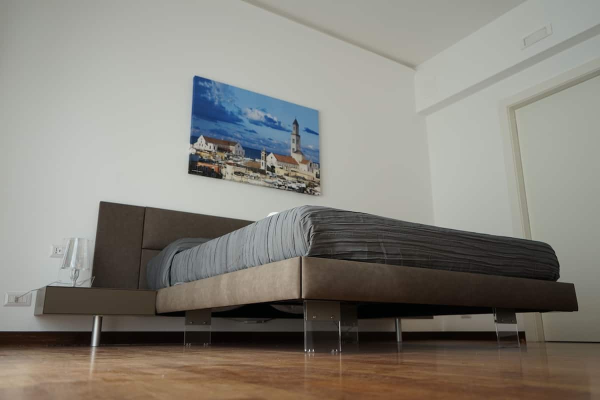 A modern bedroom with a bed on a platform, a bedside table with a lamp, and a cityscape painting on the wall.