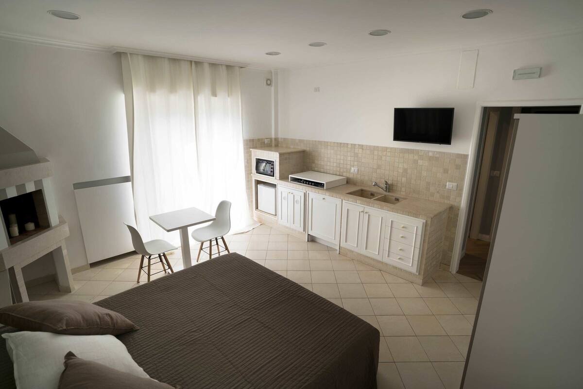 A hotel room with a bed, a desk, a chair, a kitchenette, and a TV.