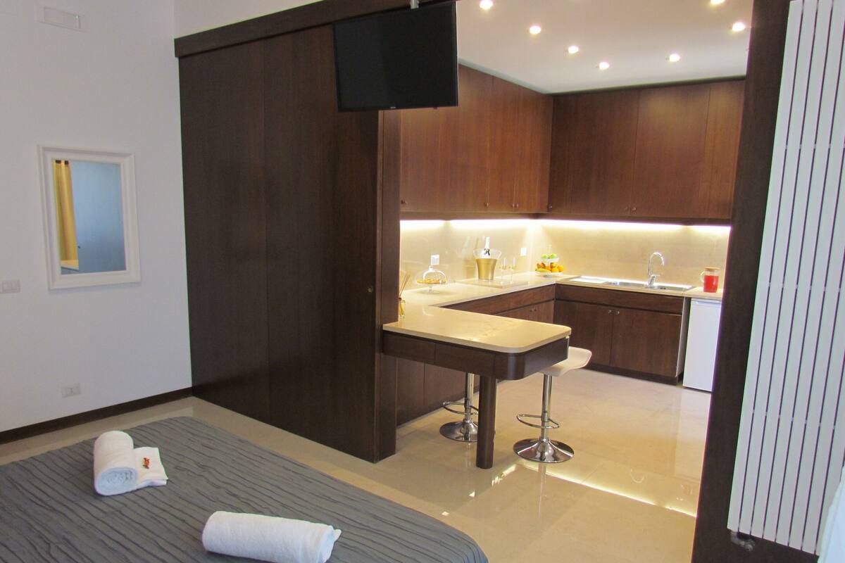 A modern hotel room with a bed, a small kitchenette, a TV, and a bathroom.