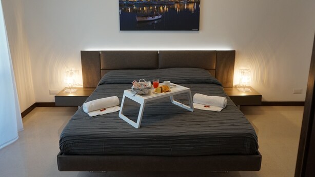A modern hotel room with a large bed, bedside tables, and a cityscape painting above. - Room 56 (Bari)