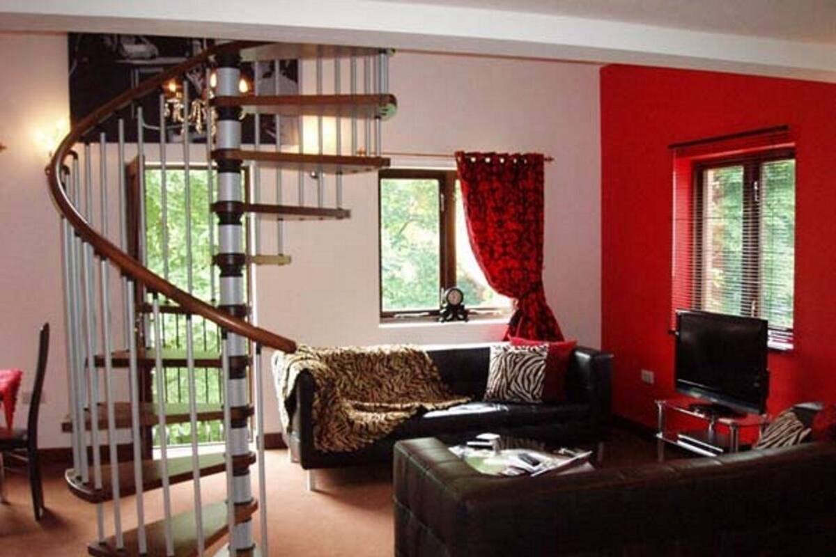 Una sala de estar con una escalera espiral, un sofá de cuero negro, un televisor y una pared roja.