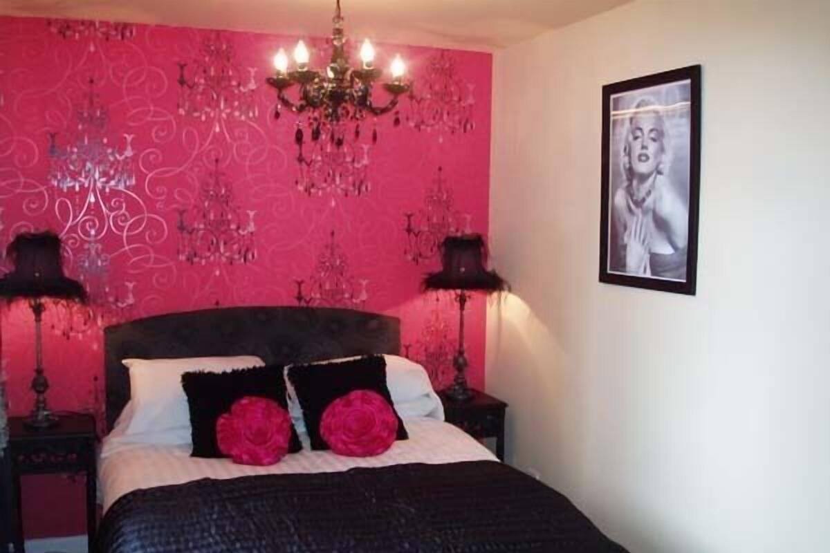 Un dormitorio con pared rosa, colcha negra y dos almohadas rojas.