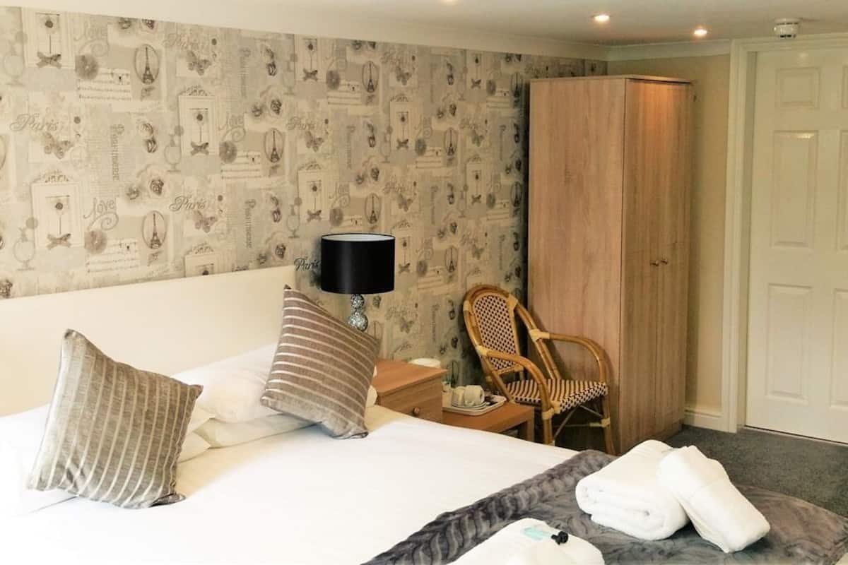 Quarto de hotel com cama, mesa de cabeceira, luminária, cadeira, guarda-roupa e porta.