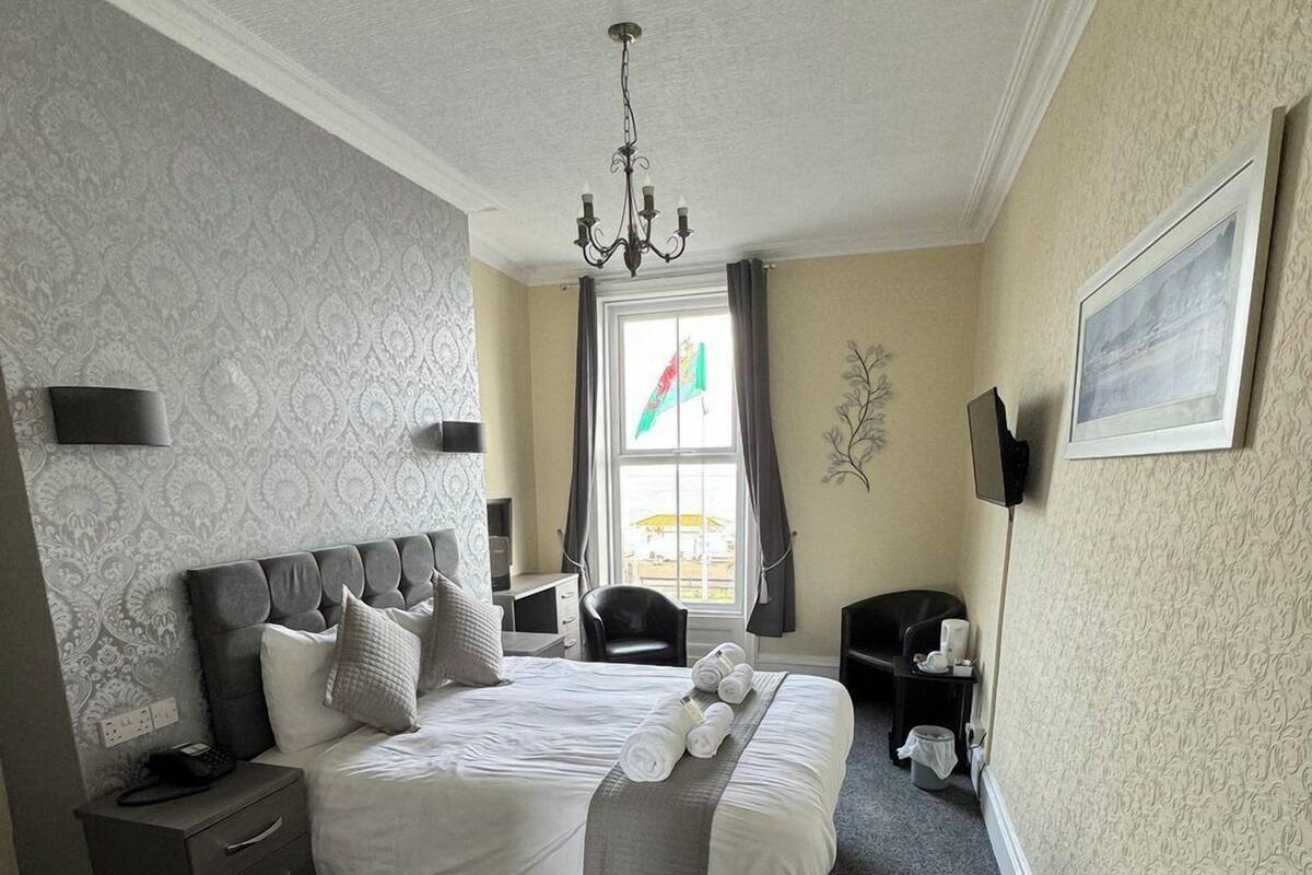 Quarto com cama, mesa de cabeceira, cadeira e janela com cortinas.