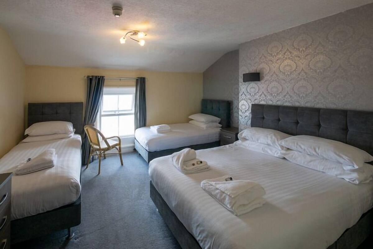 Quarto de hotel com duas camas de solteiro, uma cama de casal, uma cadeira e uma pequena mesa.