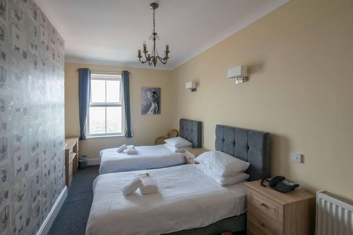 Quarto de hotel com duas camas, um candelabro, um quadro, uma janela com cortinas, um radiador e um telefone ao lado da cama.