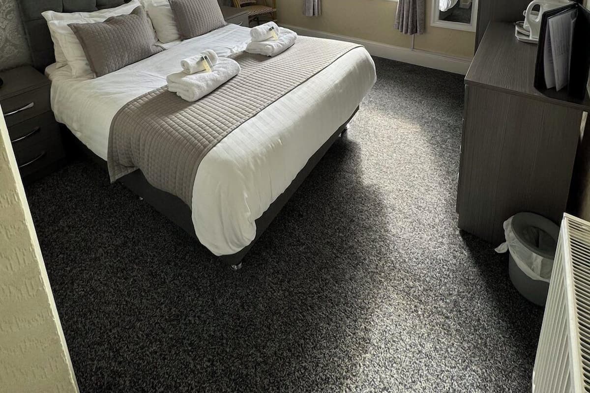 Quarto de hotel com cama, mesas de cabeceira, escrivaninha, cadeira, janela com cortinas, lustre e radiador.