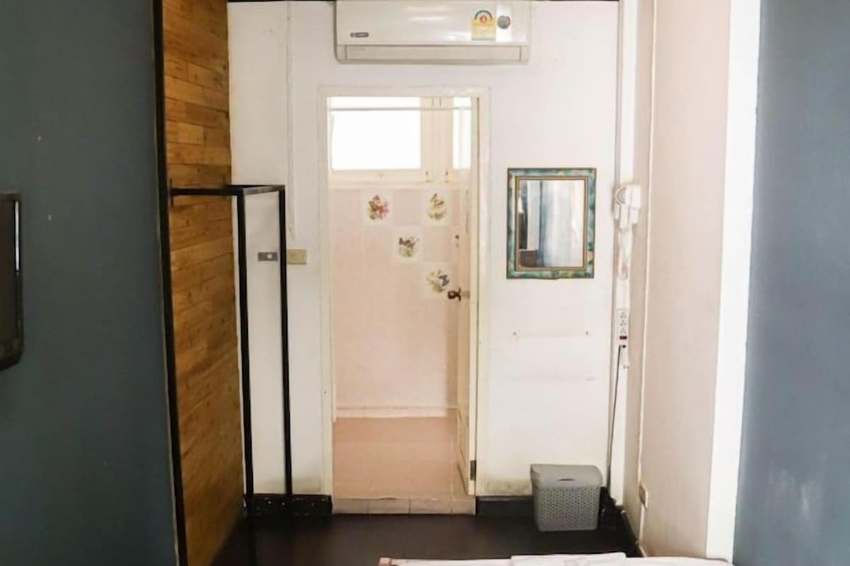 A hotel room with a bed, a wooden panel wall, an air conditioning unit, a door leading to a bathroom, and a small trash bin.