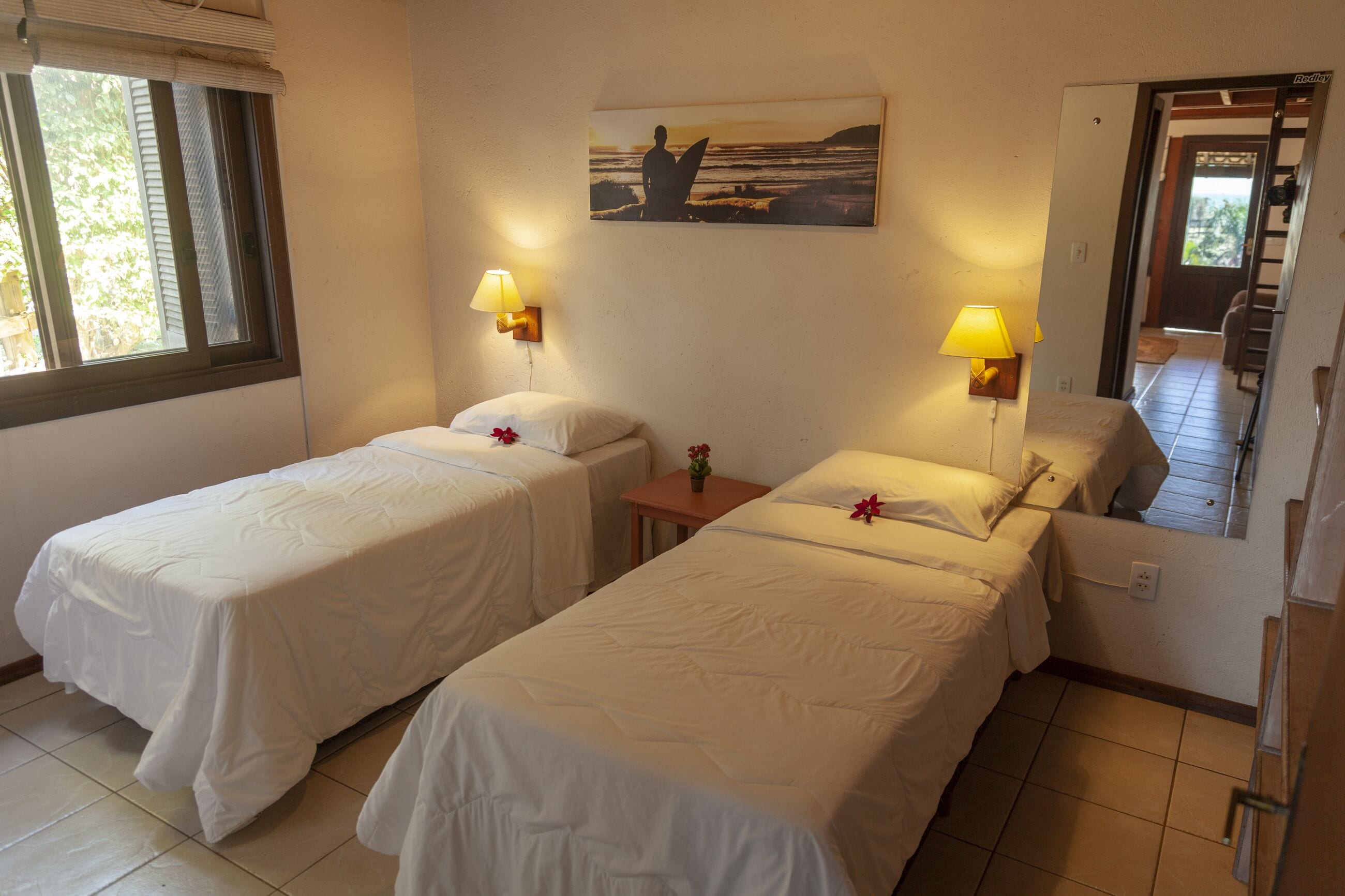 Habitación con dos camas individuales, cada una con una almohada y una mesita con una flor. Hay un cuadro en la pared sobre las camas y una ventana al lado izquierdo.