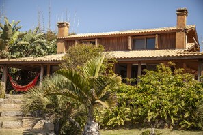A wooden house with a tiled roof, surrounded by lush greenery including palm trees and ferns.