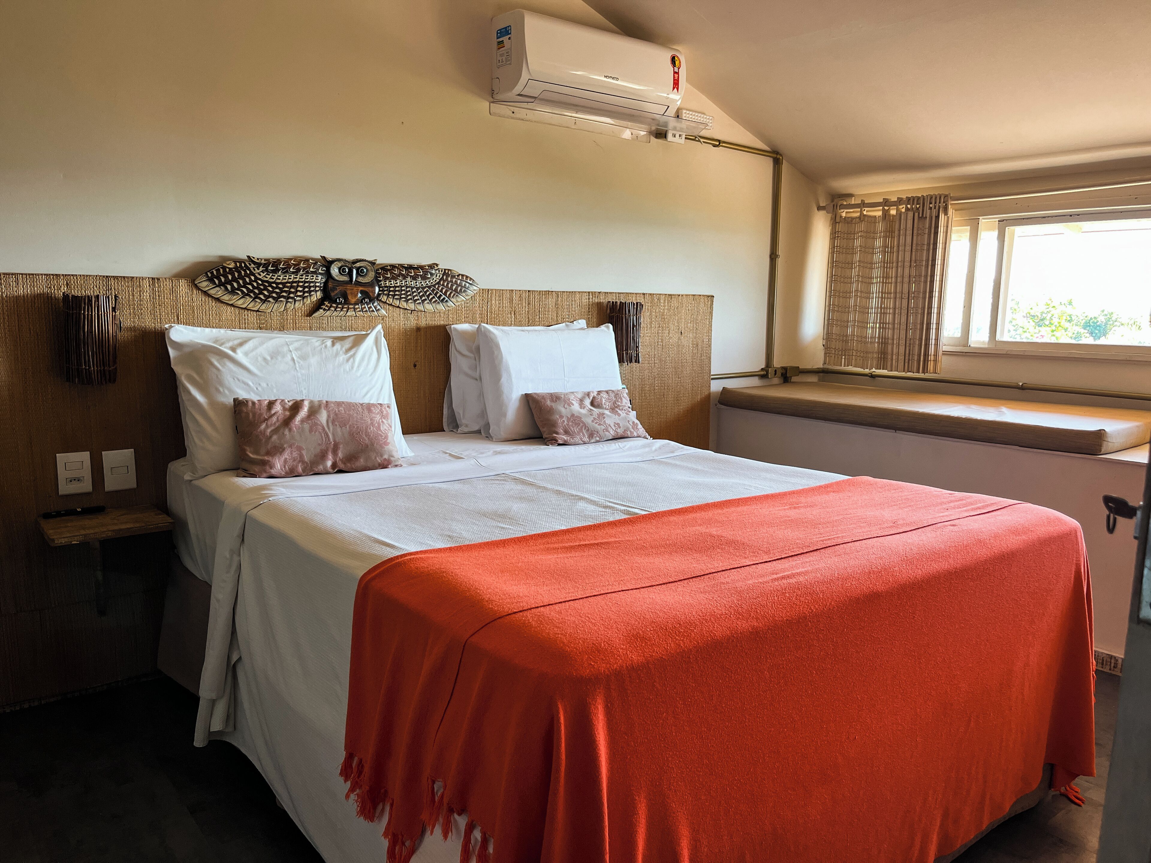 Una cama bien hecha con una colcha roja, un aire acondicionado de pared y una ventana con cortinas.
