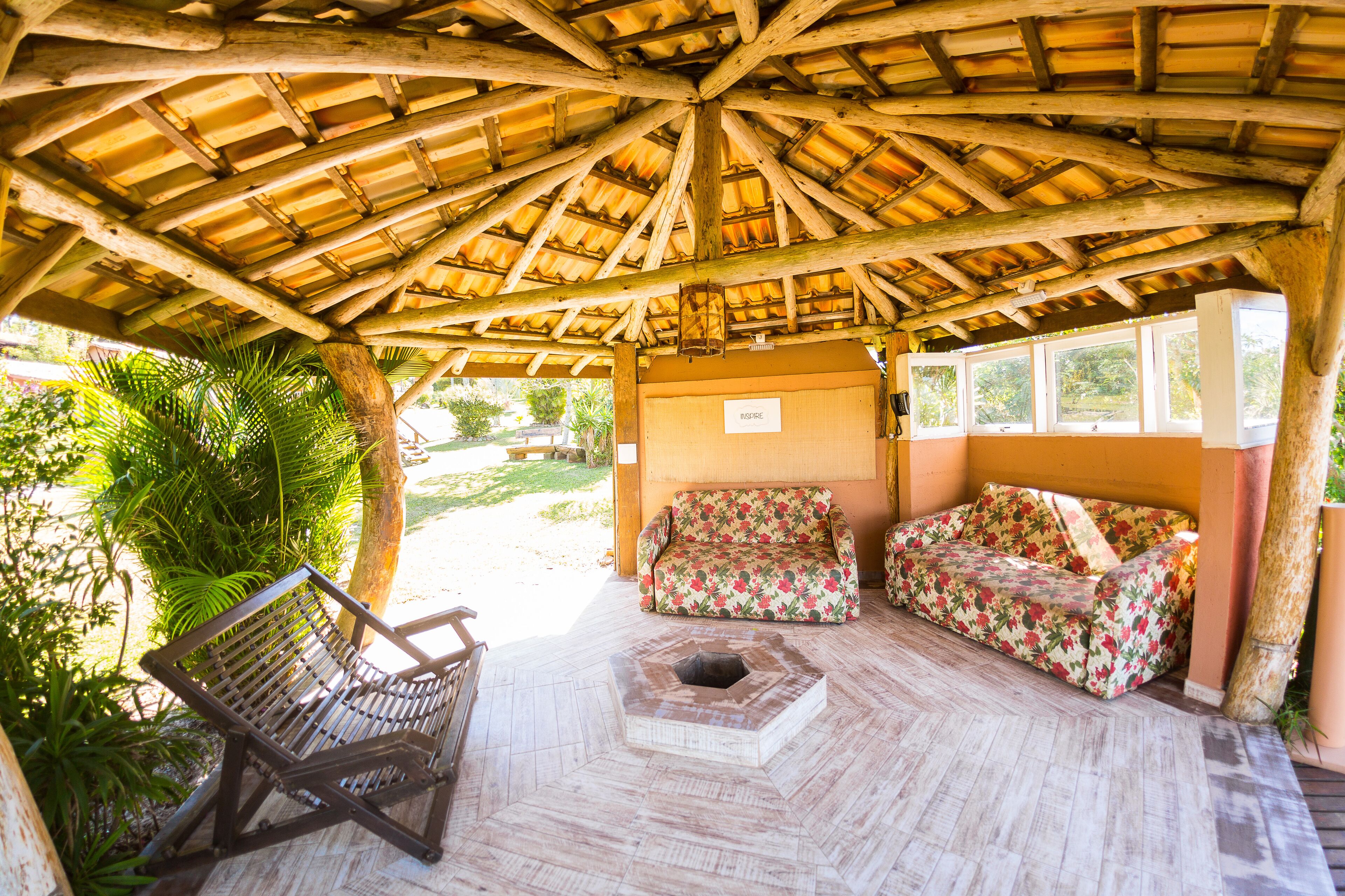 An open-air structure with a wooden roof, a bench, a sofa, and a small fire pit.