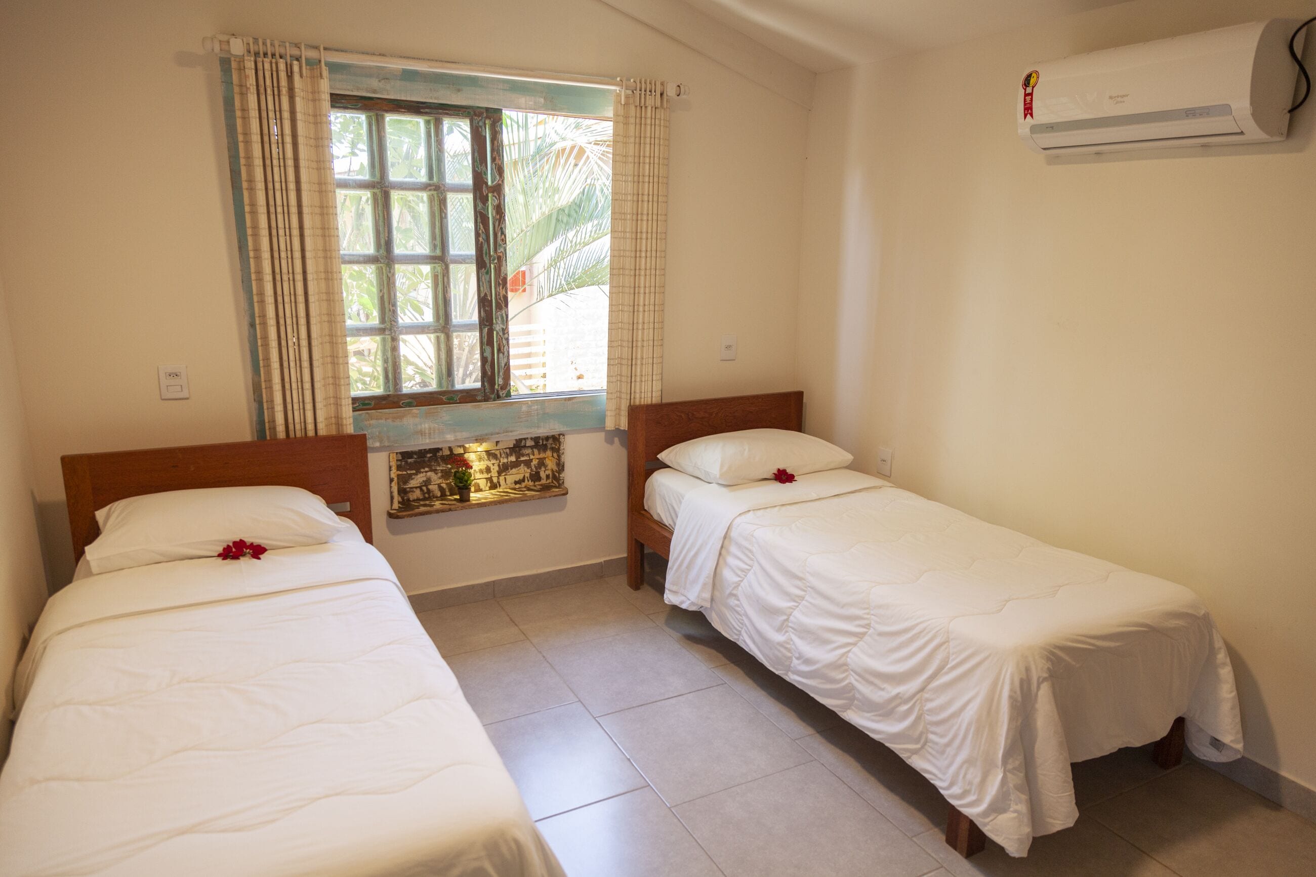 Quarto com duas camas de solteiro, janela com cortinas, ar condicionado e uma pequena decoração na parede.