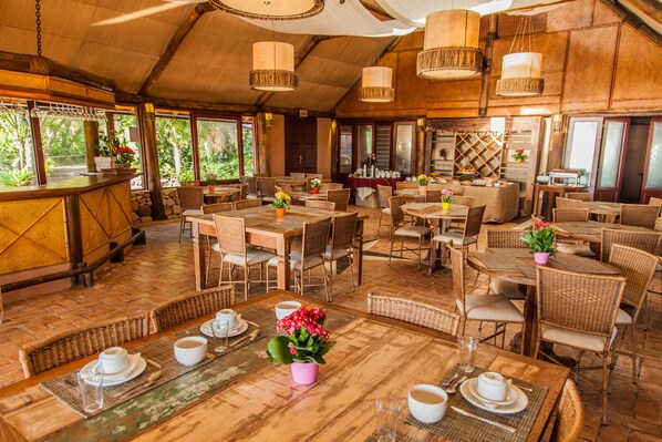An outdoor restaurant with wooden tables and chairs, potted plants, and hanging lamps.