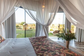 A bedroom with a large bed, sheer curtains, a view of the outdoors, and a potted plant.