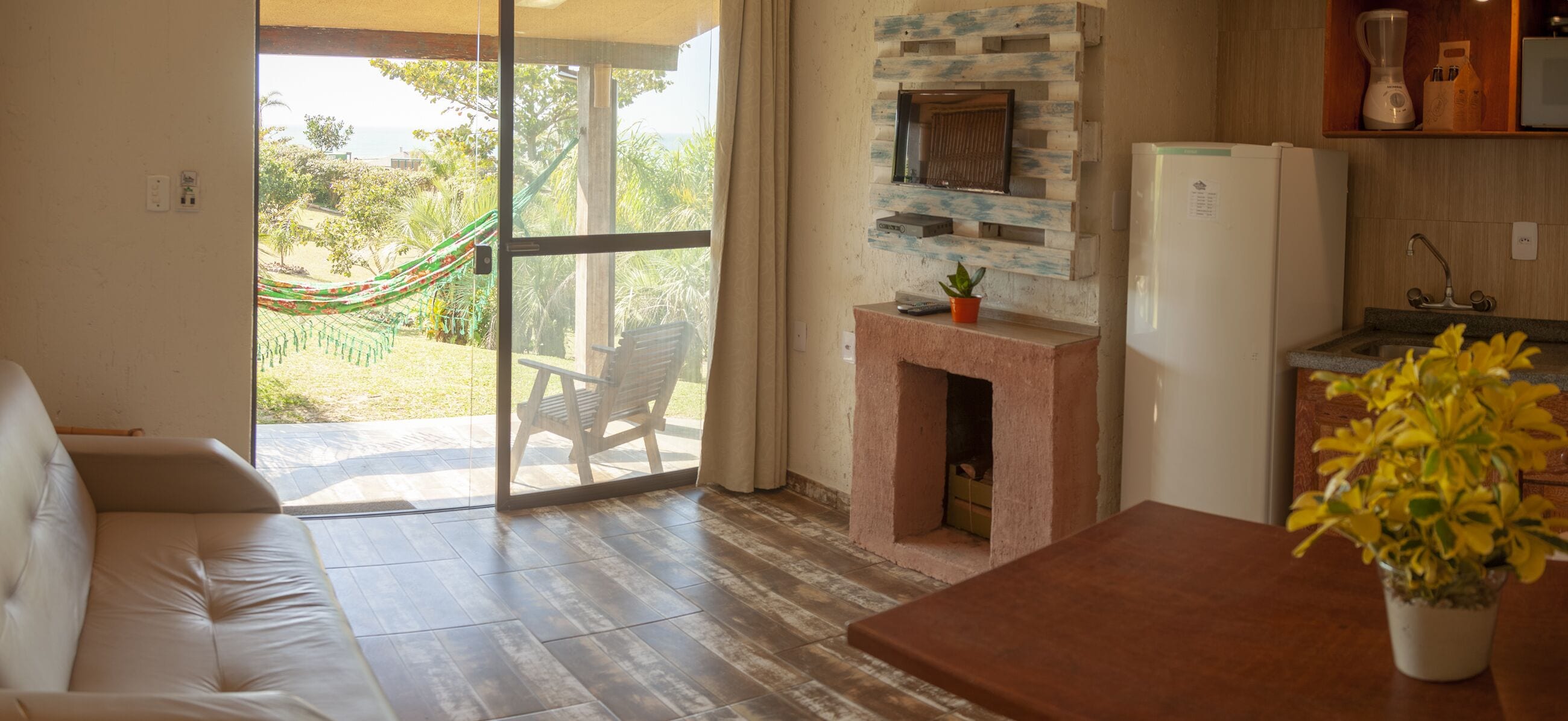 Sala de estar com sofá, lareira, cozinha com geladeira e vista para um jardim através de uma porta de vidro deslizante.