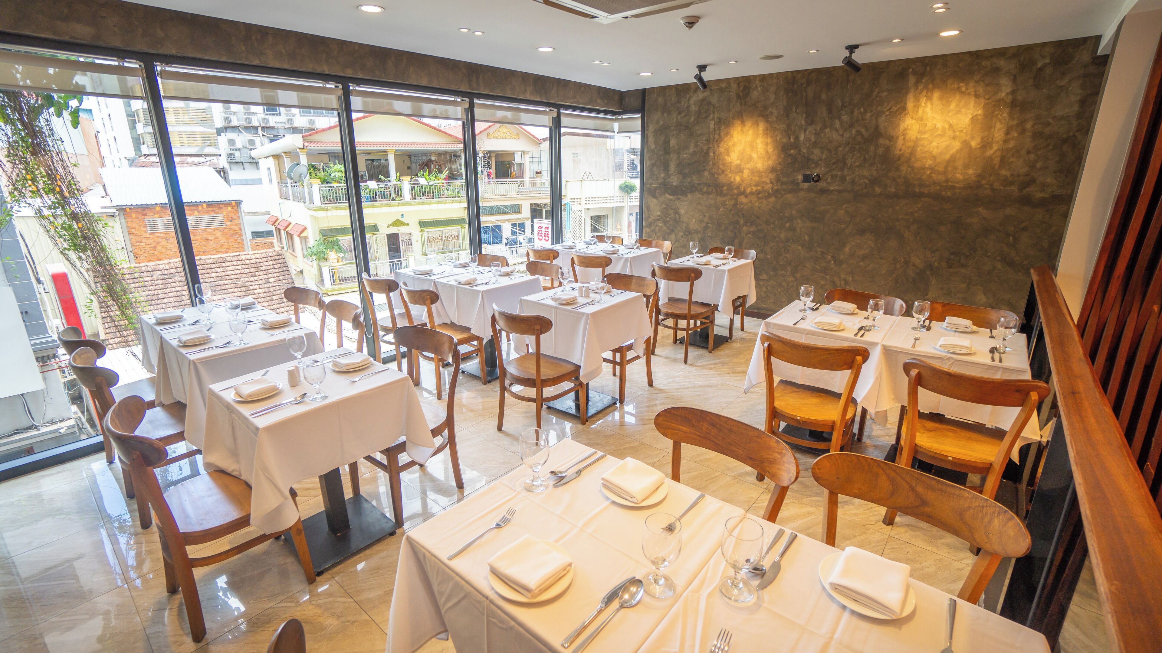 Un restaurant avec de nombreuses tables dressées pour le service, et de grandes fenêtres offrant une vue sur les bâtiments environnants.