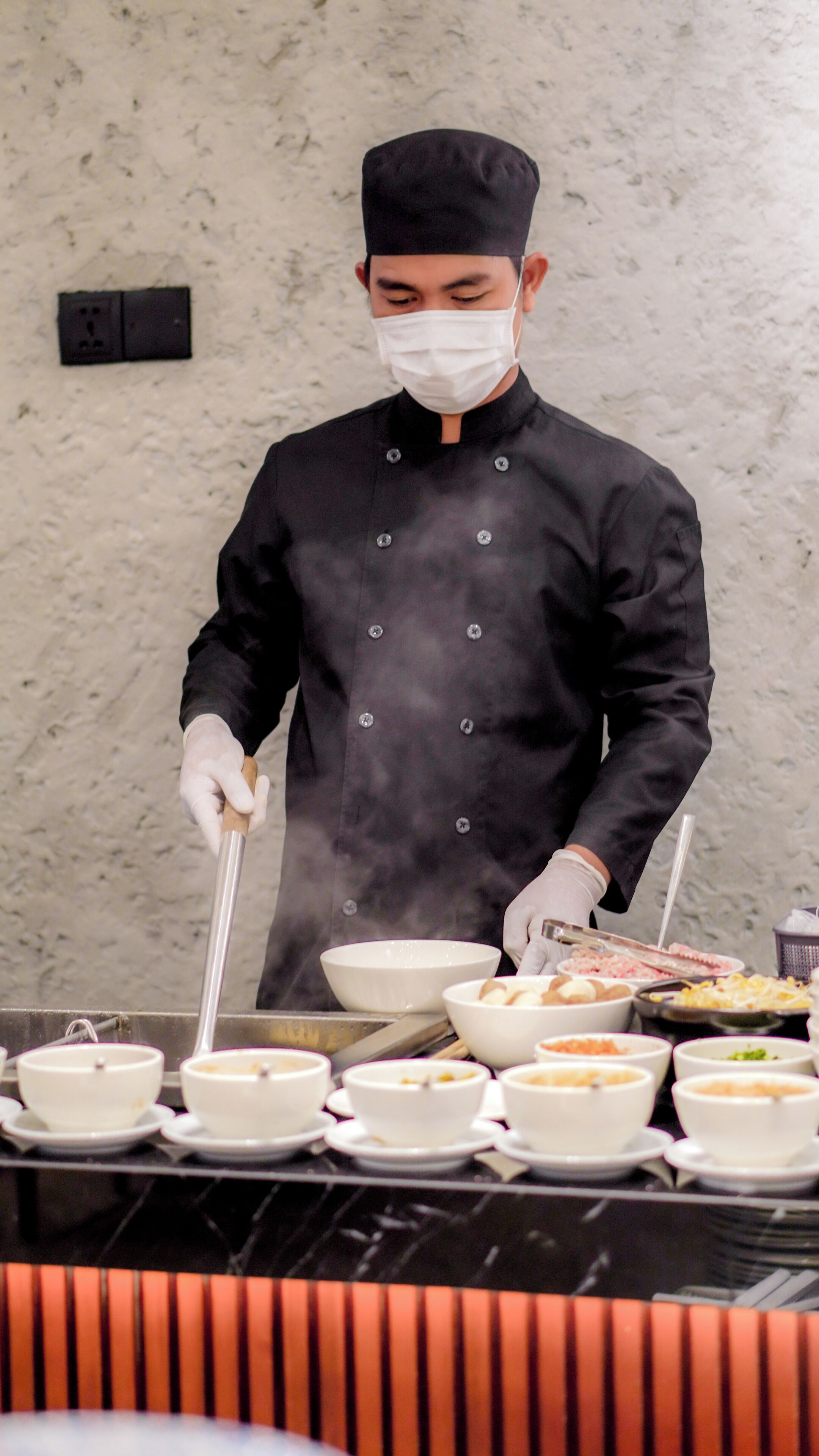 A chef in a black uniform and hat preparing food in a kitchen with multiple bowls and a grill.