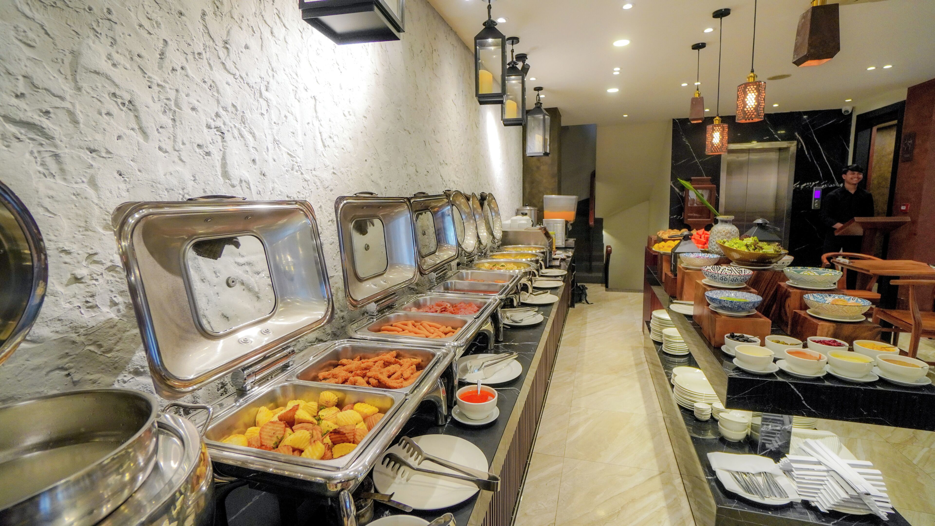 Montaje de bufé con diversas estaciones de comida, incluyendo barra de ensaladas y estación de carnes, en una sala bien iluminada con piso de mármol e iluminación moderna.
