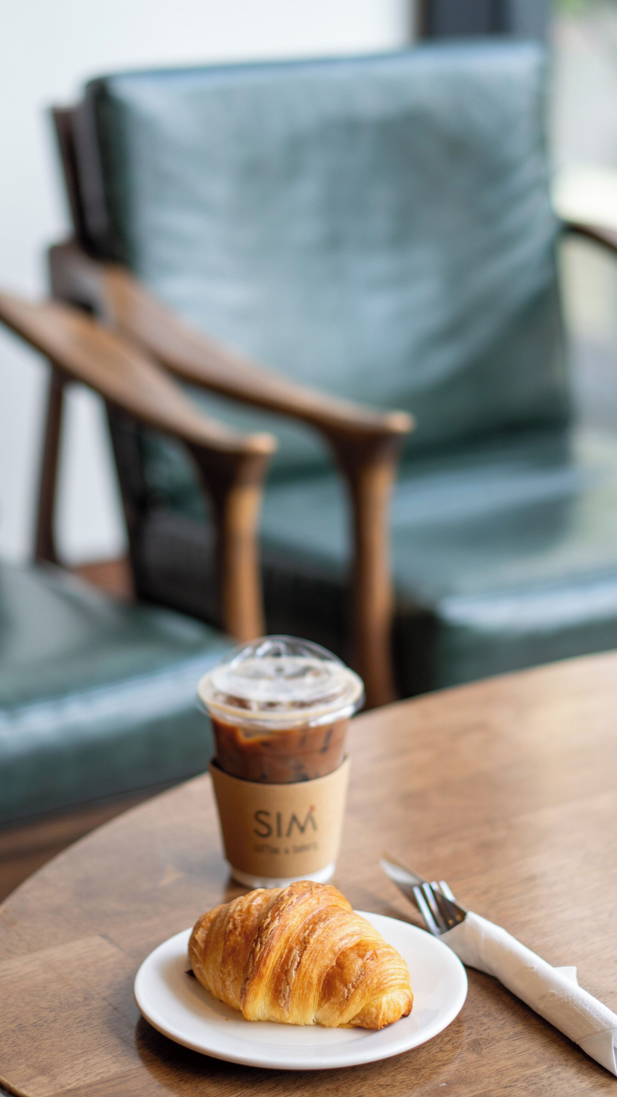 A croissant on a plate, a cup of iced coffee, and a fork on a wooden table.