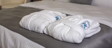 Una cama bien hecha con ropa de cama blanca y gris, un par de bata blanca con un logotipo y una lámpara al fondo.
