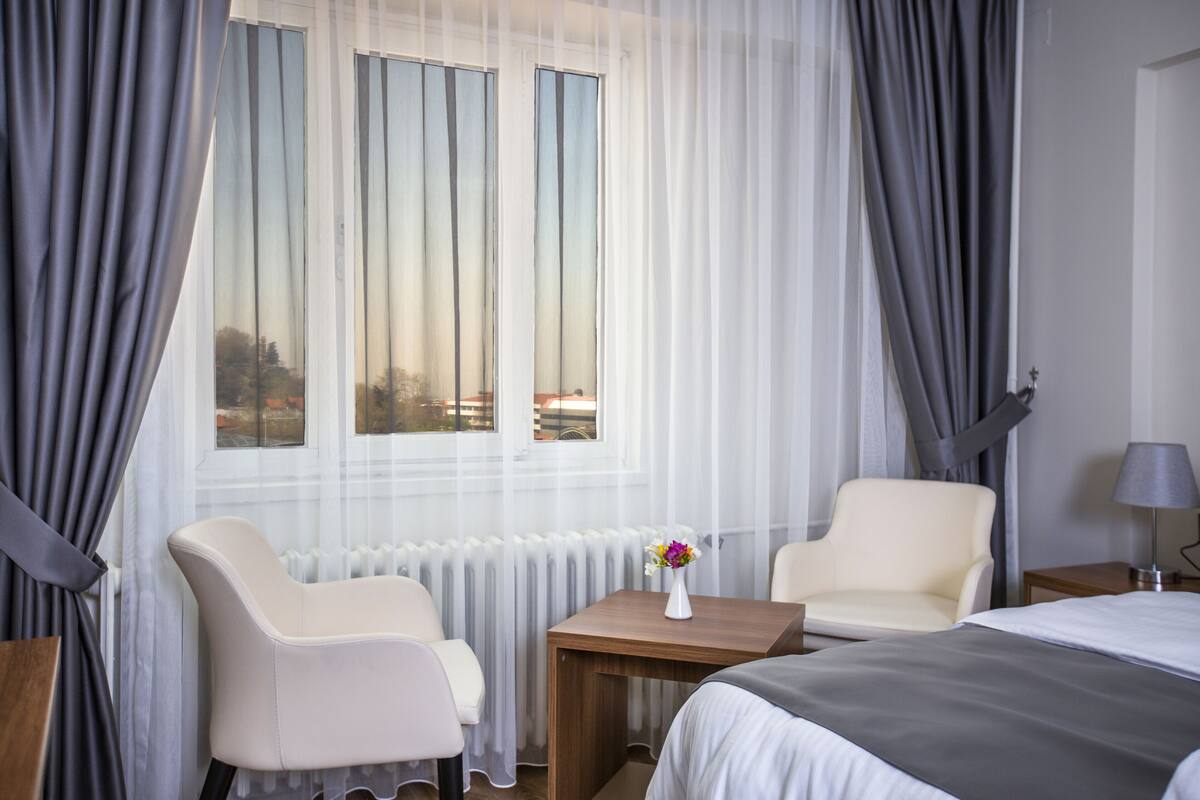 Habitación de hotel con cama, dos sillas, una mesita con un florero y una ventana con cortinas.