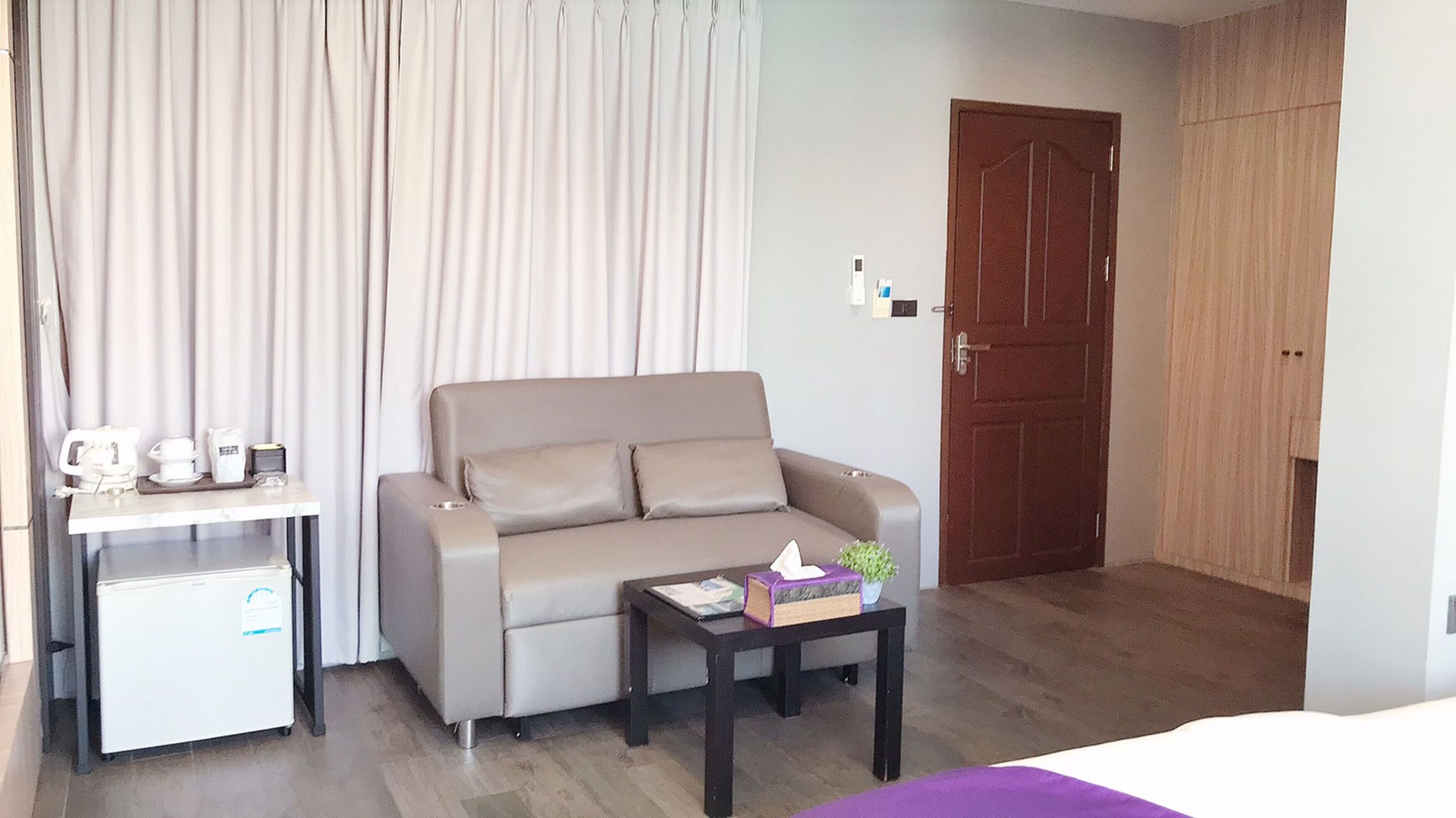 A hotel room with a sofa, a small table with a book and a plant, a coffee machine, and a mini fridge.