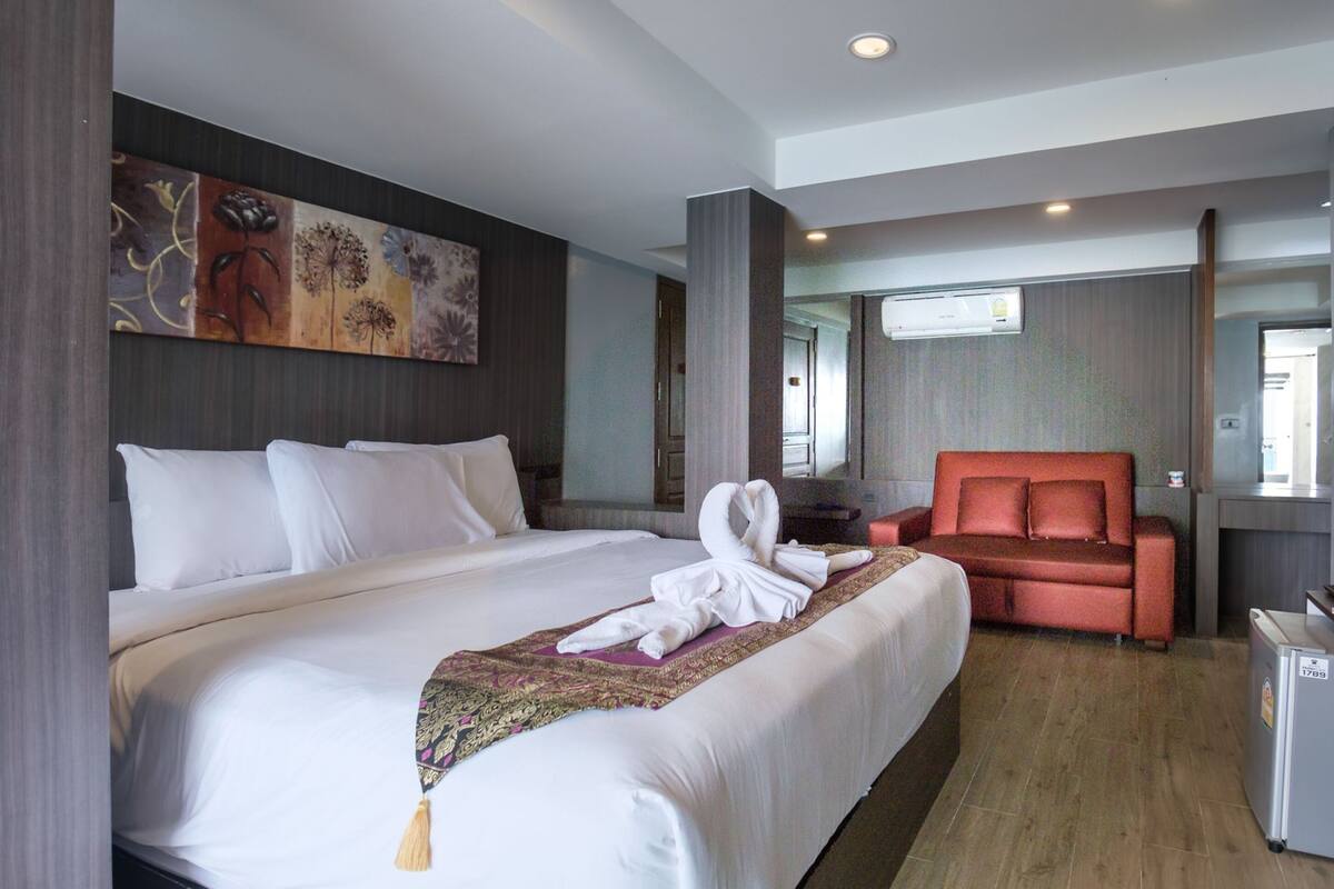 A hotel room with a large bed, a red armchair, a small desk, and a wall-mounted air conditioner.