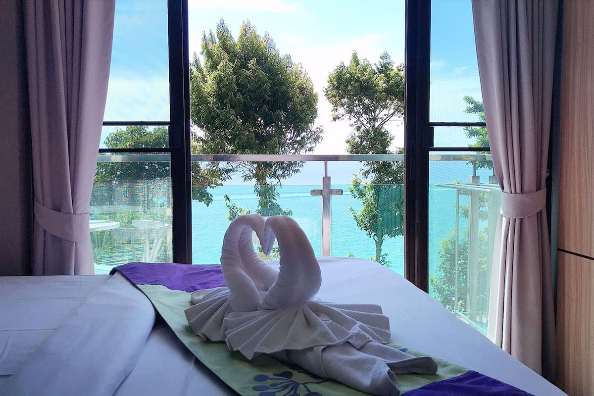 A hotel room with a bed, a window with a view of the sea and trees, and a cross on the window sill.