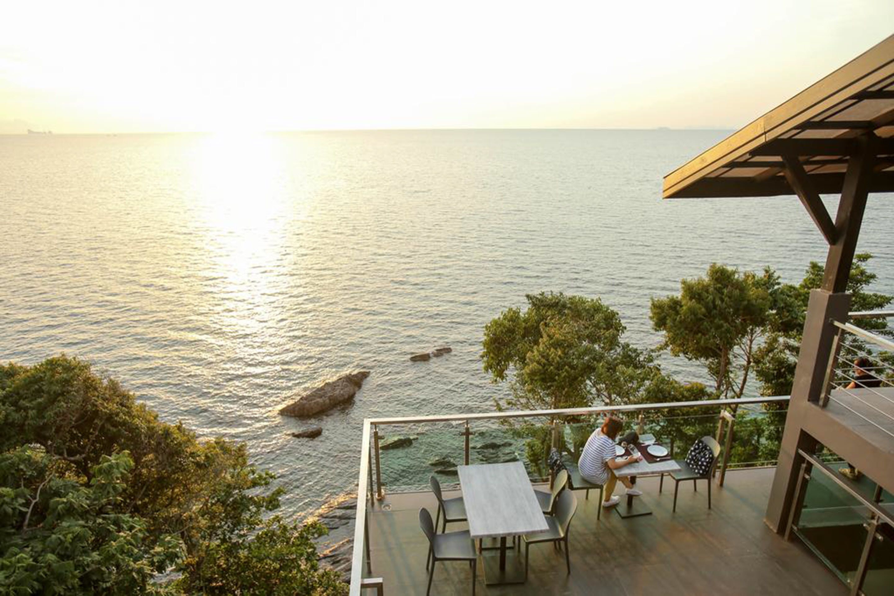 A terrace with a table and chairs overlooking a body of water, with trees and a building structure in the background.