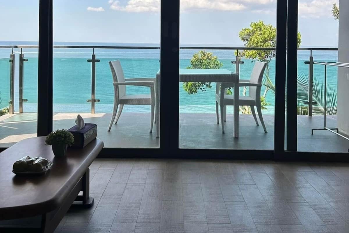 A room with a large sliding glass door leading to a balcony with a view of the ocean. There is a table and two chairs on the balcony.