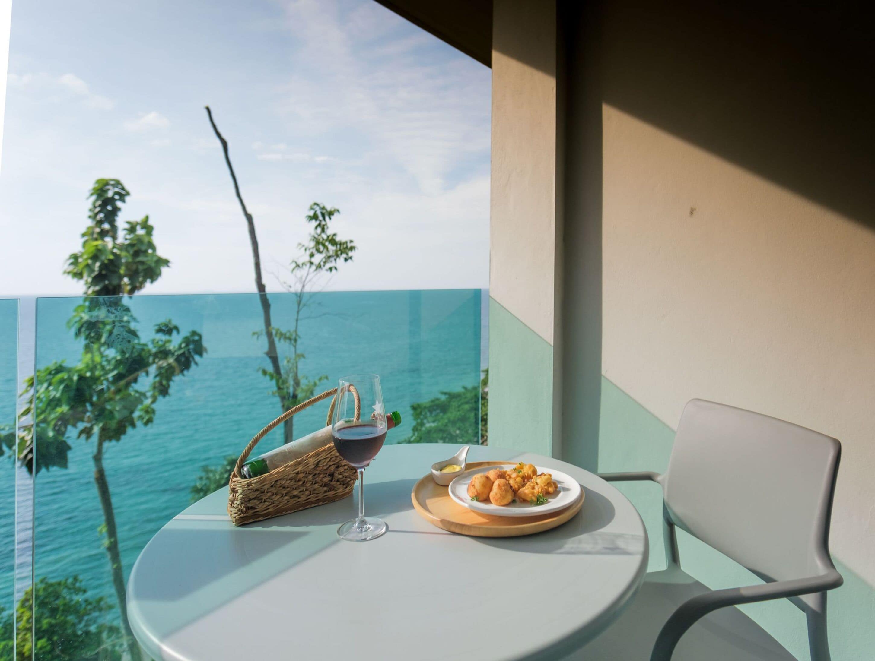 A table with a glass of red wine, a basket, and a plate of food on a balcony overlooking the sea.