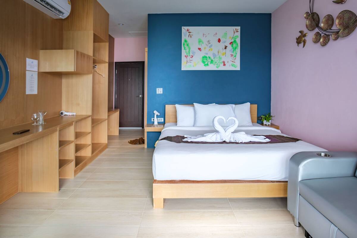 A modern hotel room with a wooden desk, a bed with a white swan-shaped pillow, a blue accent wall, and a small plant.