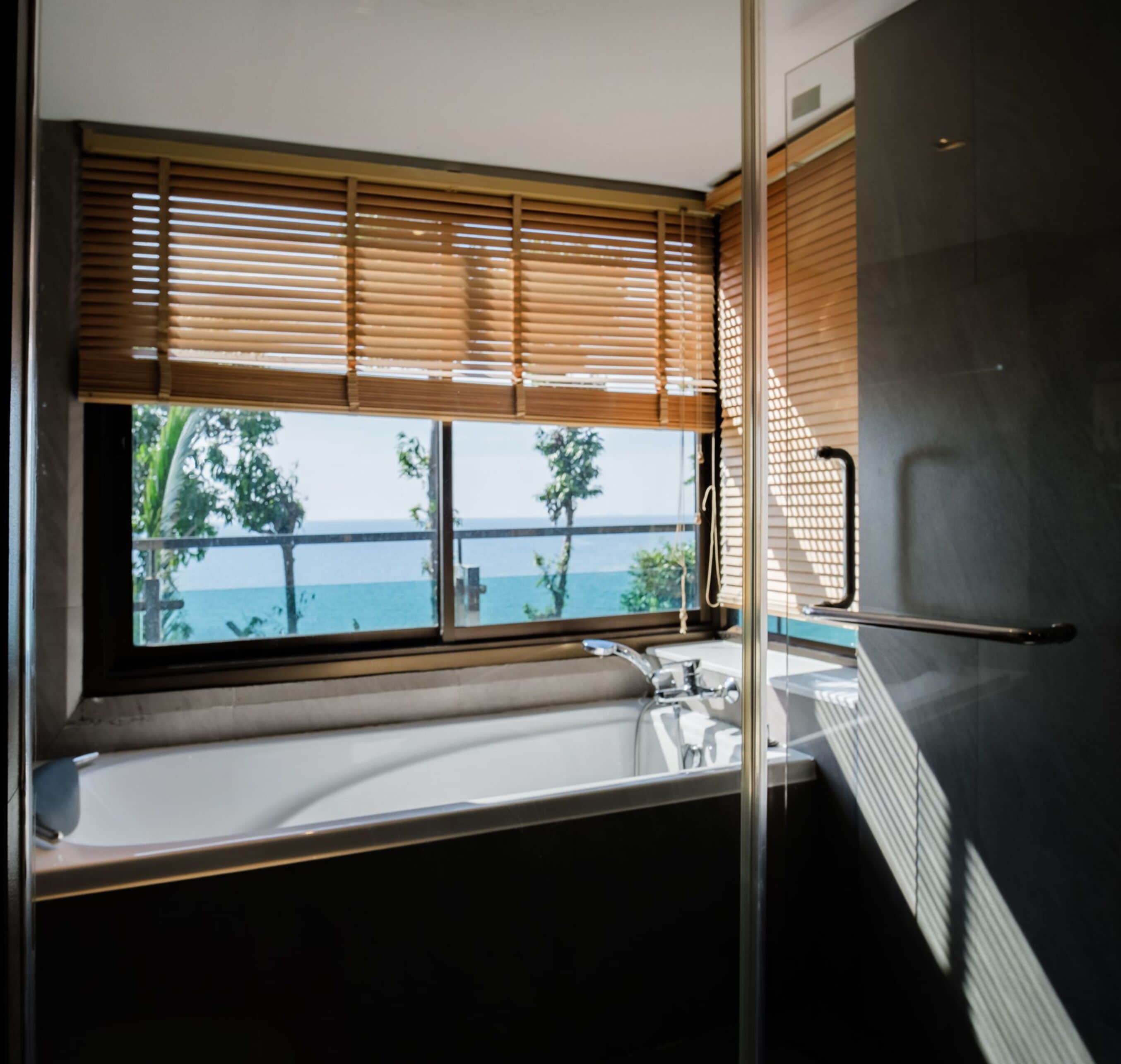 A bathroom with a bathtub, a window with wooden blinds, and a view of the sea.