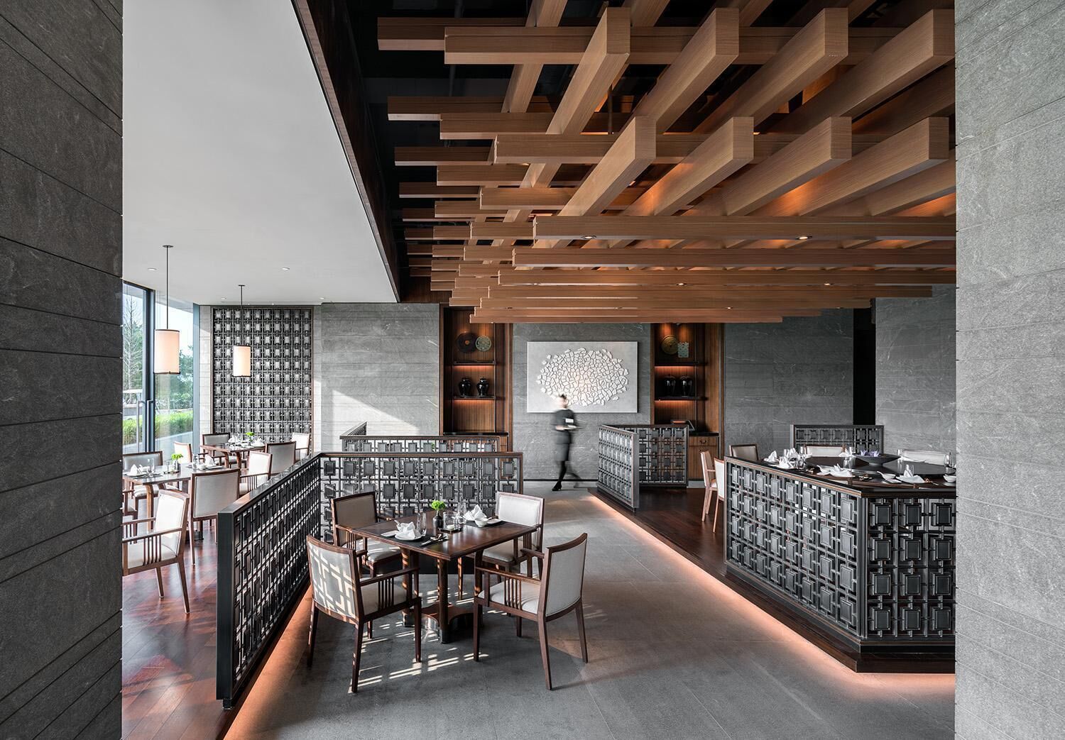 Restaurante moderno com teto alto, vigas de madeira e área de jantar central com mesas e cadeiras.