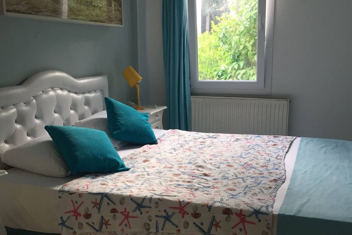 Ein Schlafzimmer mit einem weißen, gepolsterten Kopfteil, zwei türkisfarbenen Kissen, einer gemusterten Bettdecke, einem Nachttisch mit einer gelben Lampe und einem Fenster mit Blick auf Grünflächen.