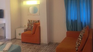 Hotellihuone, jossa on sänky, kaksi oranssia sohvaa, pieni pöytä, yöpöytä, seinään kiinnitetty lamppu ja ikkuna, jossa on siniset verhot.