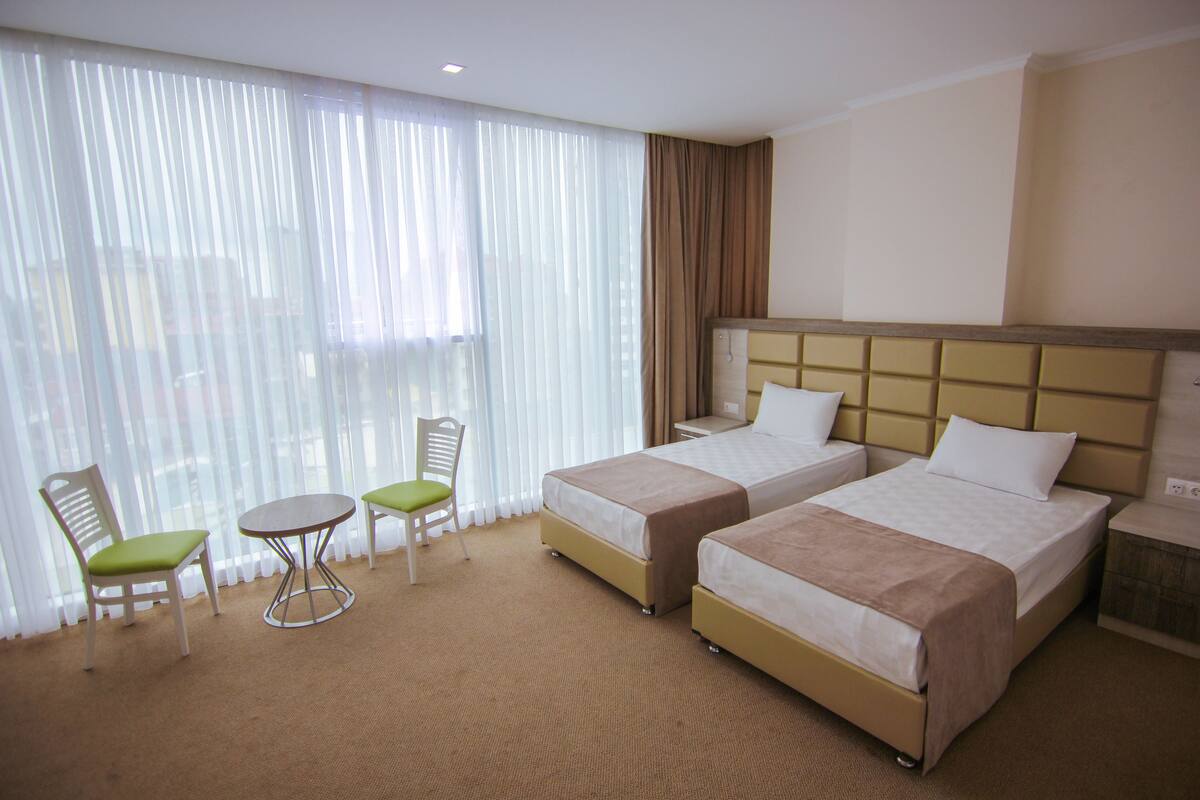 A hotel room with two beds, a small table, two chairs, and a large window with sheer curtains.