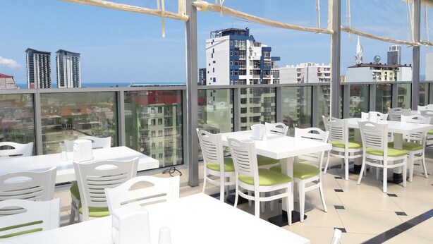 A rooftop restaurant with white tables and chairs, large windows offering a city view, and a clear sky. - Hotel Green Glass (Batumi)