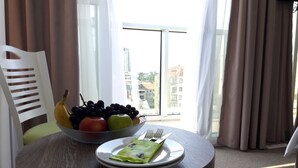 A dining area with a round table, a chair, a fruit bowl, a plate with a green packet, and a view of buildings through a window. - Hotel Green Glass (Batumi)