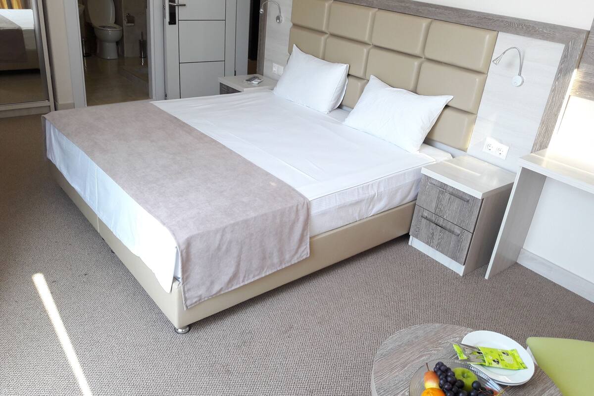 A neatly made bed with a white and beige bedspread, a bedside table with a lamp, and a fruit bowl on a small round table.