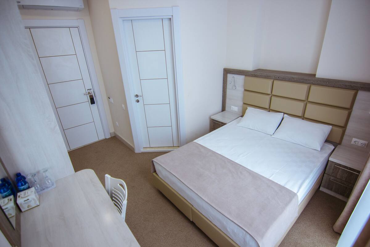 A hotel room with a bed, bedside table, and a small desk with a chair. There are two doors and a window.