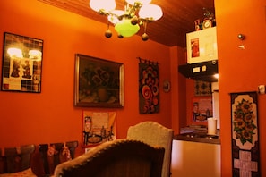 A room with orange walls, a wooden ceiling, and a chandelier. There are framed pictures and a bulletin board on the wall. - Tilly's B&B (Le Hohwald)