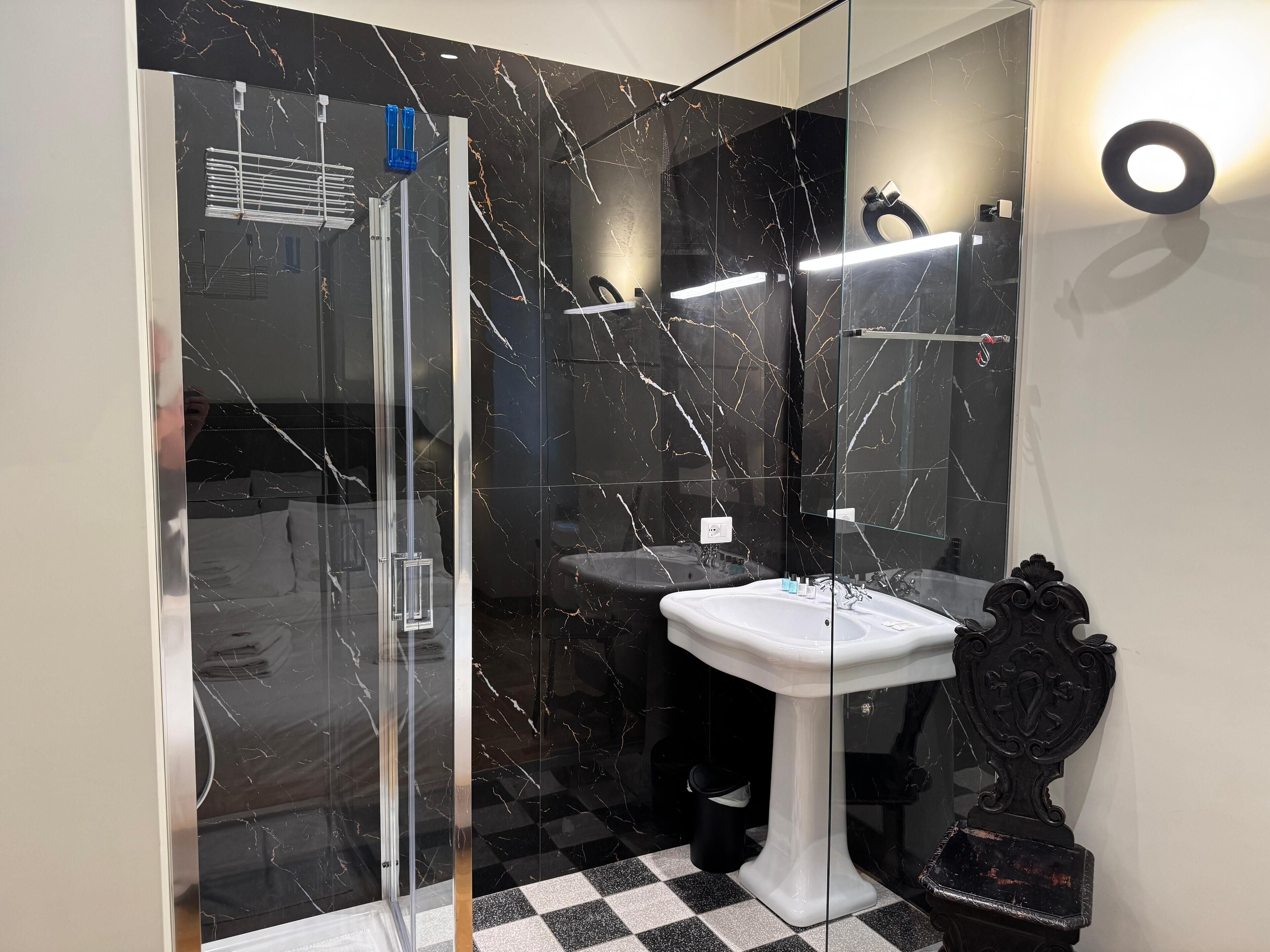 Een moderne badkamer met een douchecabine van glas, een wit wastafeltje en een zwart marmeren achterwand.