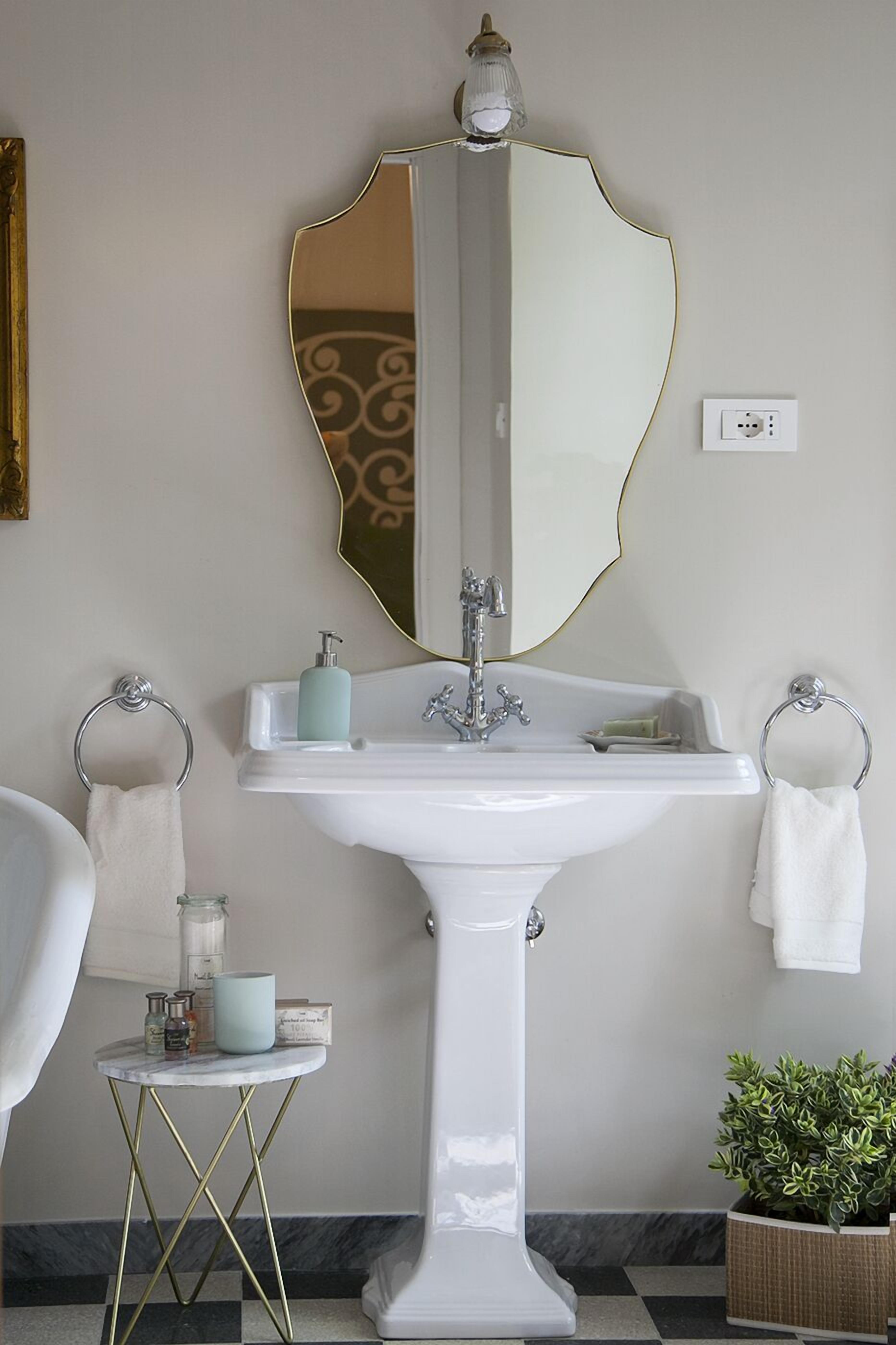 A bathroom with a white pedestal sink, a large mirror with a gold frame, and a small round table with toiletries.