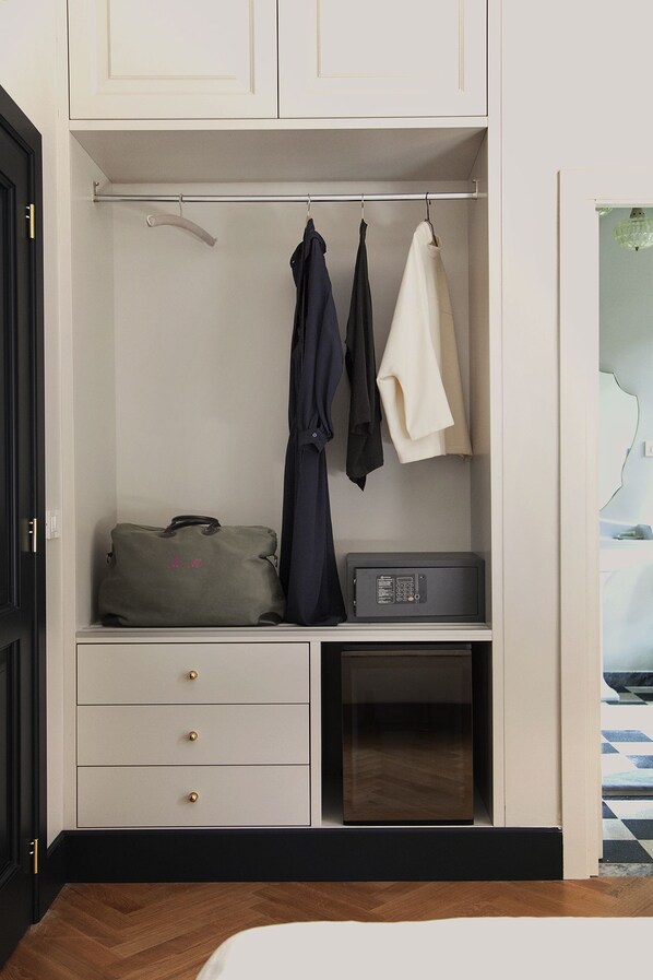 A hotel room with a wardrobe containing a suitcase, a coat, and a handbag. There is a small table with a radio and a drawer with three compartments.